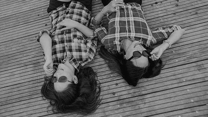 A greyscale top-view image of two young women lying down on the floor wearing checked shirts and sunglasses.
