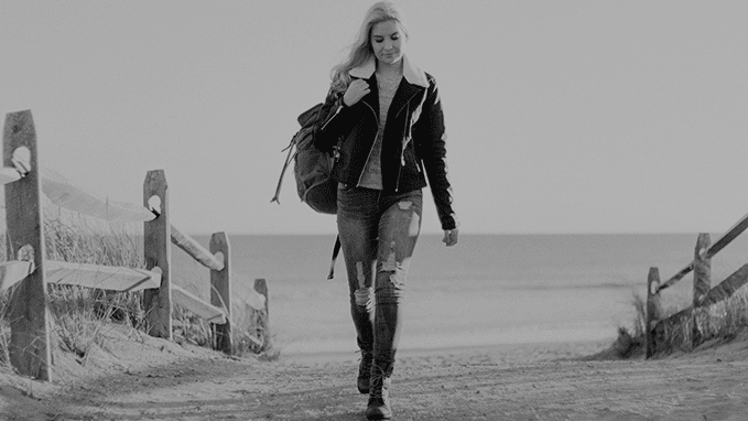 A greyscale image of a woman walking in a sandy area with a backpack.